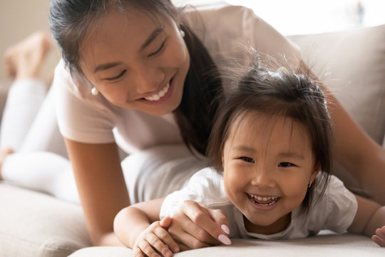 Close up image, Asian toddler kid girl spending active funny time with mother or babysitter at home. Lying on couch in living room mom laughing relish weekend activity with adorable little daughter