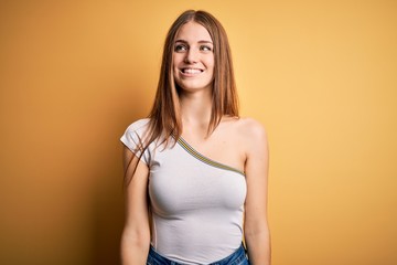 Obraz premium Young beautiful redhead woman wearing casual t-shirt over isolated yellow background looking away to side with smile on face, natural expression. Laughing confident.