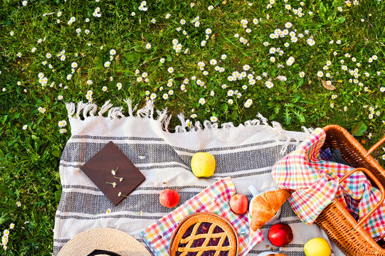 Picnic basket on a plaid and a green meadow with flowers.Lunch sweet cake, croissants, drinks, fruits in the park on the green grass. Summer picnic background concept. Above, Copy space