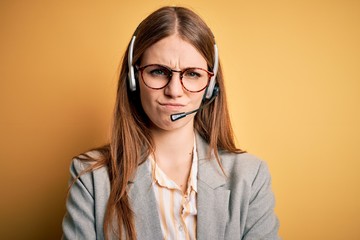 Young redhead call center agent woman overworked wearing glasses using headset skeptic and nervous,...