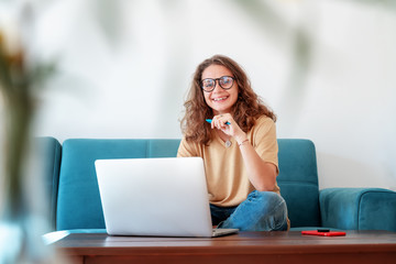 Fototapeta na wymiar Charming beautiful young girl in glasses with curly hair sits on a blue sofa at home in front of a laptop, remote work and education, beauty and fashion.