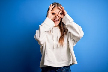 Young beautiful redhead sporty woman wearing sweatshirt over isolated blue background doing ok gesture like binoculars sticking tongue out, eyes looking through fingers. Crazy expression.