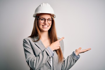 Young beautiful redhead architect woman wearing security helmet over white background amazed and smiling to the camera while presenting with hand and pointing with finger.