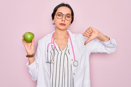Young beautiful doctor woman with blue eyes wearing stethoscope holding green apple fruit with angry face, negative sign showing dislike with thumbs down, rejection concept