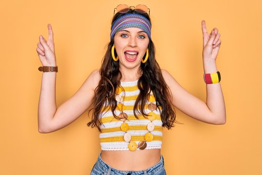 Young beautiful hippie woman with blue eyes wearing accesories and sunnglasses smiling amazed and surprised and pointing up with fingers and raised arms.