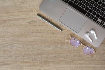 Lifestyle picture. Inspiring and modern workspace. Wooden table with  grey laptop, glasses, metallic pen and white wireless headphones. Space on the left for text. Cosy style.Top view, flat lay.