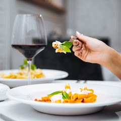 Female hand eating penne pasta with mozzarella cheeze with a glass of red wine in an italian restaurant, square format