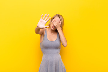 young blonde woman covering face with hand and putting other hand up front to stop camera, refusing...
