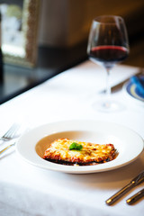 Appetizing fresh cooked italian cuisine dish lasagna with a glass of red wine on the white plate on the white table, vertical format
