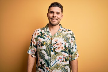 Young man with blue eyes on vacation wearing floral summer shirt over yellow background with a...