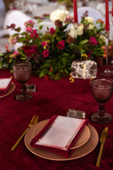 Place at the banquet table: the menu card lies on a red napkin on plates, a gilt fork and knife on the sides of the plate, a glass of brown glass, flower arrangement, red candles