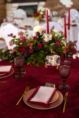 Place at the banquet table: the menu card lies on a red napkin on plates, a gilt fork and knife on the sides of the plate, a glass of brown glass, flower arrangement, red candles
