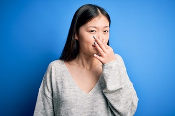 Fototapeta na wymiar Young beautiful asian woman wearing casual sweater standing over blue isolated background smelling something stinky and disgusting, intolerable smell, holding breath with fingers on nose. Bad smell