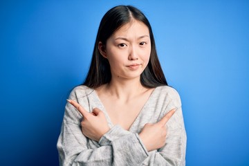 Young beautiful asian woman wearing casual sweater standing over blue isolated background Pointing to both sides with fingers, different direction disagree
