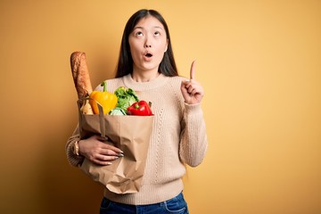 Young asian woman holding paper bag of fresh healthy groceries over yellow isolated background amazed and surprised looking up and pointing with fingers and raised arms.