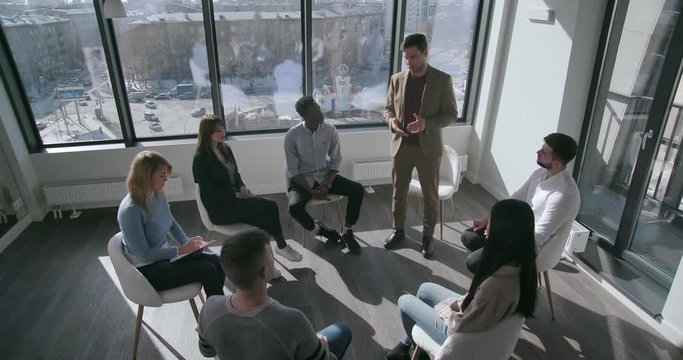 Top view of an experienced young male psychologist in formal clothes tells mixed race audience how to deal with fears and phobias while hiring in the sunny conference hall
