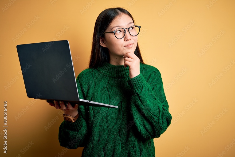 Poster Young asian business woman wearing glasses and working using computer laptop with hand on chin thinking about question, pensive expression. Smiling with thoughtful face. Doubt concept.