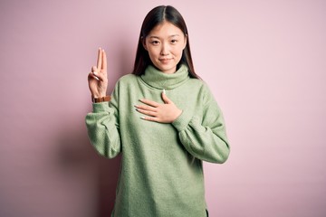 Young beautiful asian woman wearing green winter sweater over pink solated background smiling swearing with hand on chest and fingers up, making a loyalty promise oath