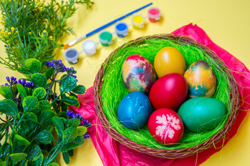 Green grass nest in a basket with colored chicken Easter eggs, multicolored painted eggs,paints and plastic flowers, Easter tradition, celebration concept 