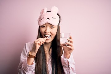 Young beautiful asian woman wearing pajama and sleep mask brushing her teeth using tooth brush, oral paste and glass of water, cleaning teeth and tongue as healthy health care morning routine