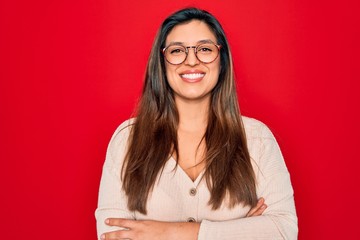 Young hispanic smart woman wearing glasses standing over red isolated background happy face smiling with crossed arms looking at the camera. Positive person.