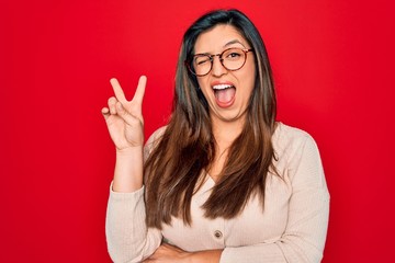 Young hispanic smart woman wearing glasses standing over red isolated background smiling with happy face winking at the camera doing victory sign. Number two.