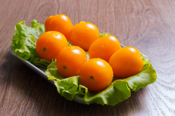 Yellow cherry tomatoes covered with drops of water on a lettuce leaf lie on a plate. Plate is on a wooden tabletop.