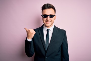 Young handsome caucasian business man wearing funny thug life glasses smiling with happy face looking and pointing to the side with thumb up.