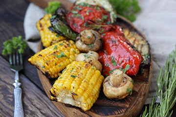 Grilled vegetables with herbs on a wooden board on a wooden background. Roasted corn, pepper, mushrooms, zucchini, eggplant with rosemary and parsley with fork. Rustic. Picnic. Background image
