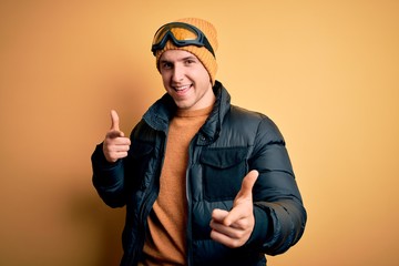 Young handsome caucasian man wearing hat, coat and ski glasses for winter and snow weather pointing fingers to camera with happy and funny face. Good energy and vibes.