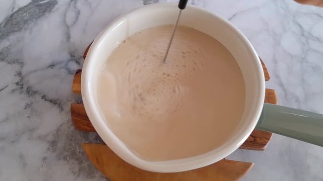 Frothing plant milk for morning latte coffee in an enamel saucepan with electric frother. Top view