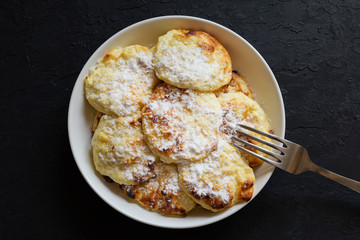 Sweet cottage cheese pancakes on a white plate sprinkled with powdered sugar with a fork on a dark background. Syrniki, curd fritters baked in the oven. Easy to prepare homemade breakfast. Top view.