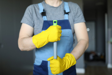 Close-up of professional cleaner holding mop. Male in special blue uniform and yellow protective gloves for work. Detergents for cleanliness. Cleaning service and renovation concept