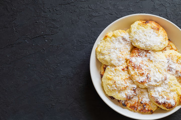 Sweet cottage cheese pancakes on a dark background. Syrniki, curd fritters baked in the oven. Pancakes with cottage cheese on a white plate sprinkled with powdered sugar. Copy space.