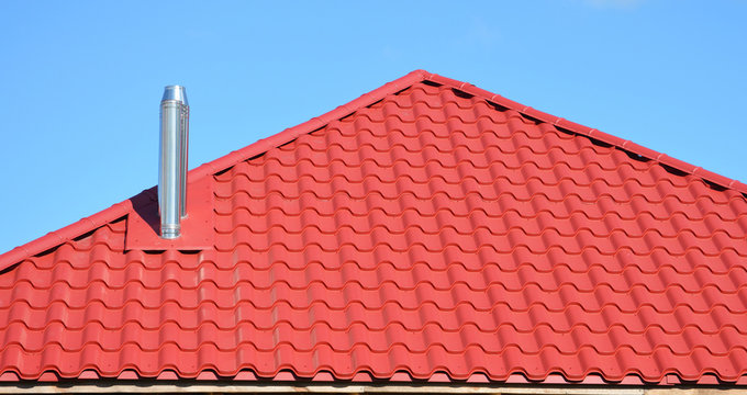 Roofing construction covered with red metal roof tiles and steel chimney installed against blue sky.