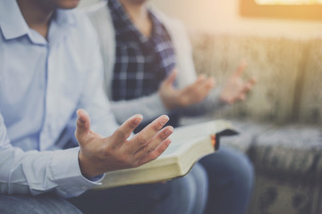 Close up of a man holding a bible and raise hand up  praying to God with his friends while sitting on the sofa at home, Christian family worship concept, copy space