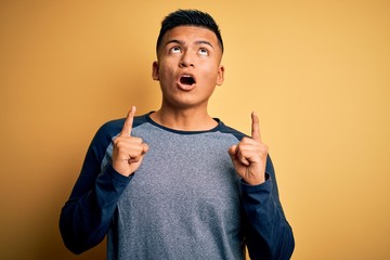 Young handsome latin man wearing casual t-shirt standing over yellow background amazed and surprised looking up and pointing with fingers and raised arms.