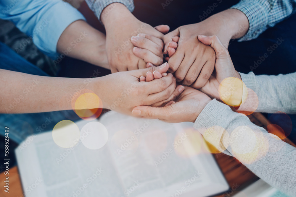 Wall mural close up of people group holding hand and pray together over a blurred holy bible on wooden table, c