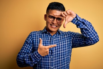 Young handsome latin man wearing casual shirt and glasses over yellow background smiling making frame with hands and fingers with happy face. Creativity and photography concept.