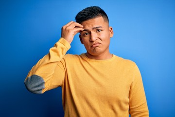 Young handsome latin man wearing yellow casual sweater over isolated blue background worried and stressed about a problem with hand on forehead, nervous and anxious for crisis