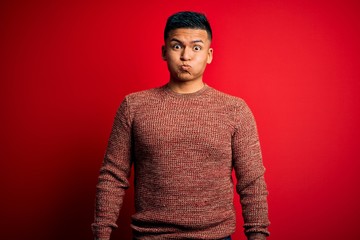 Young handsome latin man wearing casual sweater standing over red background puffing cheeks with funny face. Mouth inflated with air, crazy expression.