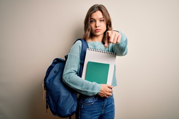 Young blonde student girl wearing backpack and books from school over isolated background pointing with finger to the camera and to you, hand sign, positive and confident gesture from the front