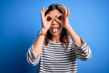 Young beautiful blonde girl wearing casual sweater standing over blue isolated background doing ok gesture like binoculars sticking tongue out, eyes looking through fingers. Crazy expression.