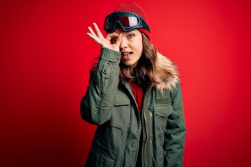 Young blonde girl wearing ski glasses and winter coat for ski weather over red background doing ok gesture with hand smiling, eye looking through fingers with happy face.