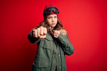 Young blonde girl wearing ski glasses and winter coat for ski weather over red background Punching fist to fight, aggressive and angry attack, threat and violence