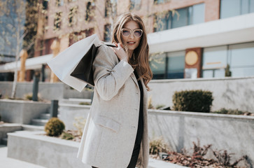 Attractive cute girl with the shopping bags and warm drink in her hand. Modern city megapolis on background. Lady order and shop online. Beautiful caucasian lady