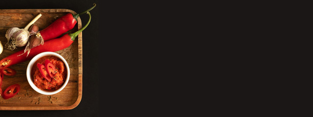 Harissa sauce in the gravy boat. Ingredients for cooking - chili peppers, garlic and spices. Everything lies on a wooden tray. Black background, copy space and top view