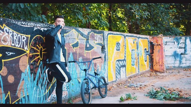 Handsome confident man is standing on graffiti background near BMX bike in a skatepark. Copy space. 4K.