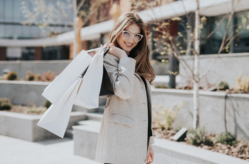 Fototapeta premium Attractive cute girl with the shopping bags and warm drink in her hand. Modern city megapolis on background. Lady order and shop online. Beautiful caucasian lady
