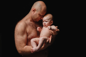 father holds a newborn son in his arms. Bald man with a naked torso and a 4 month old baby on a black background